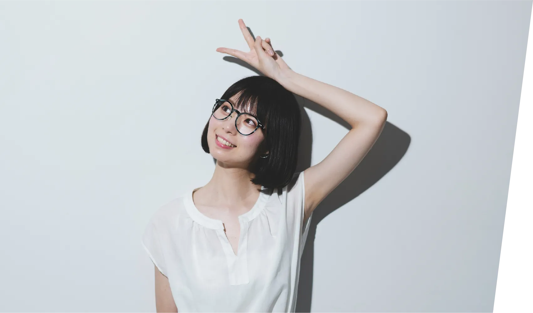 丸みのある黒縁のメガネをかけた女性が斜め右上を向き、ピースサインをする様子